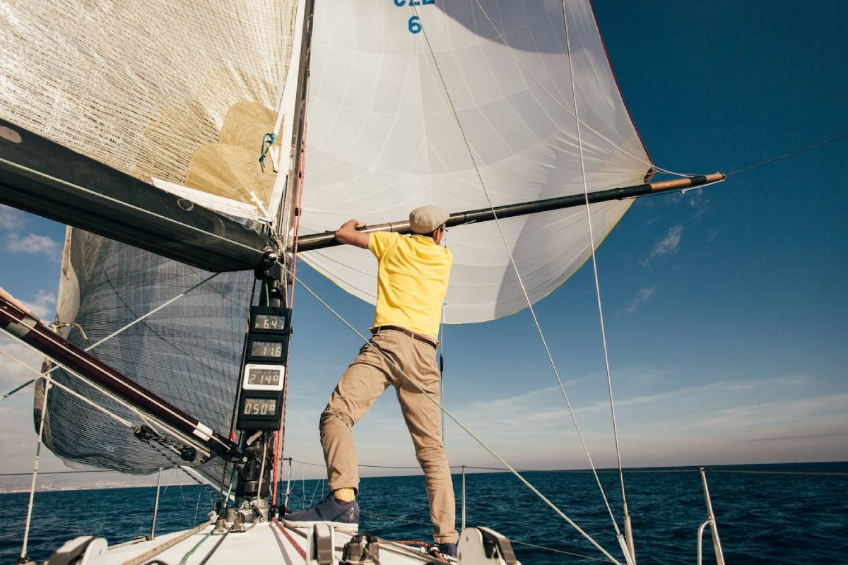 A sailor in a yellow shirt adjusts the sail on a yacht, navigating the blue sea under a clear sky. 