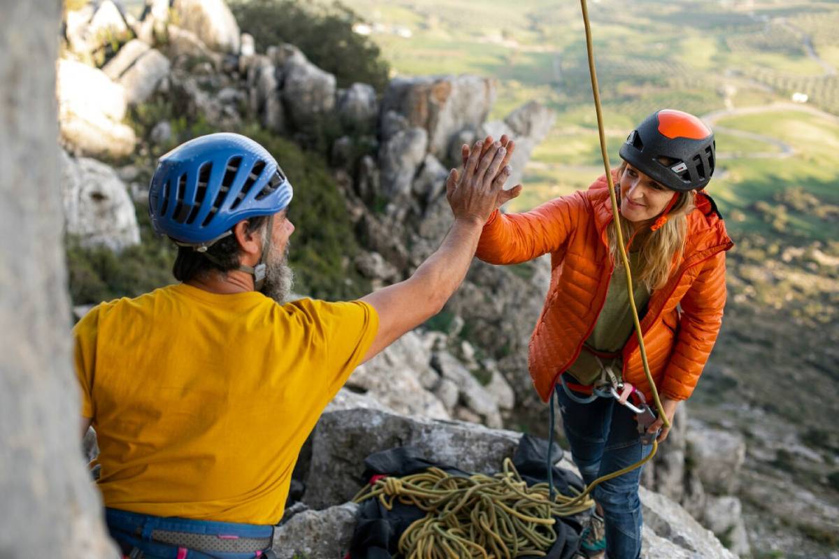  A climber in a bright orange jacket exchanges a high-five with a partner on a rocky cliff, surrounded by greenery and distant hills.