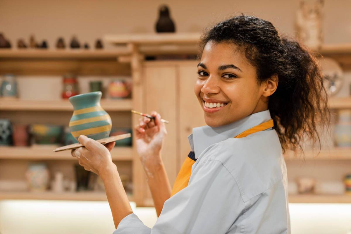  A person holds a painted ceramic pot in one hand, using a brush with the other, surrounded by art supplies and decorated pottery in the background.
