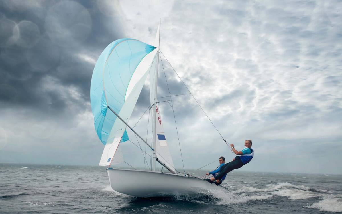 Two sailors maneuver a sailboat with a bright blue spinnaker under a dramatic, cloudy sky on choppy waters.