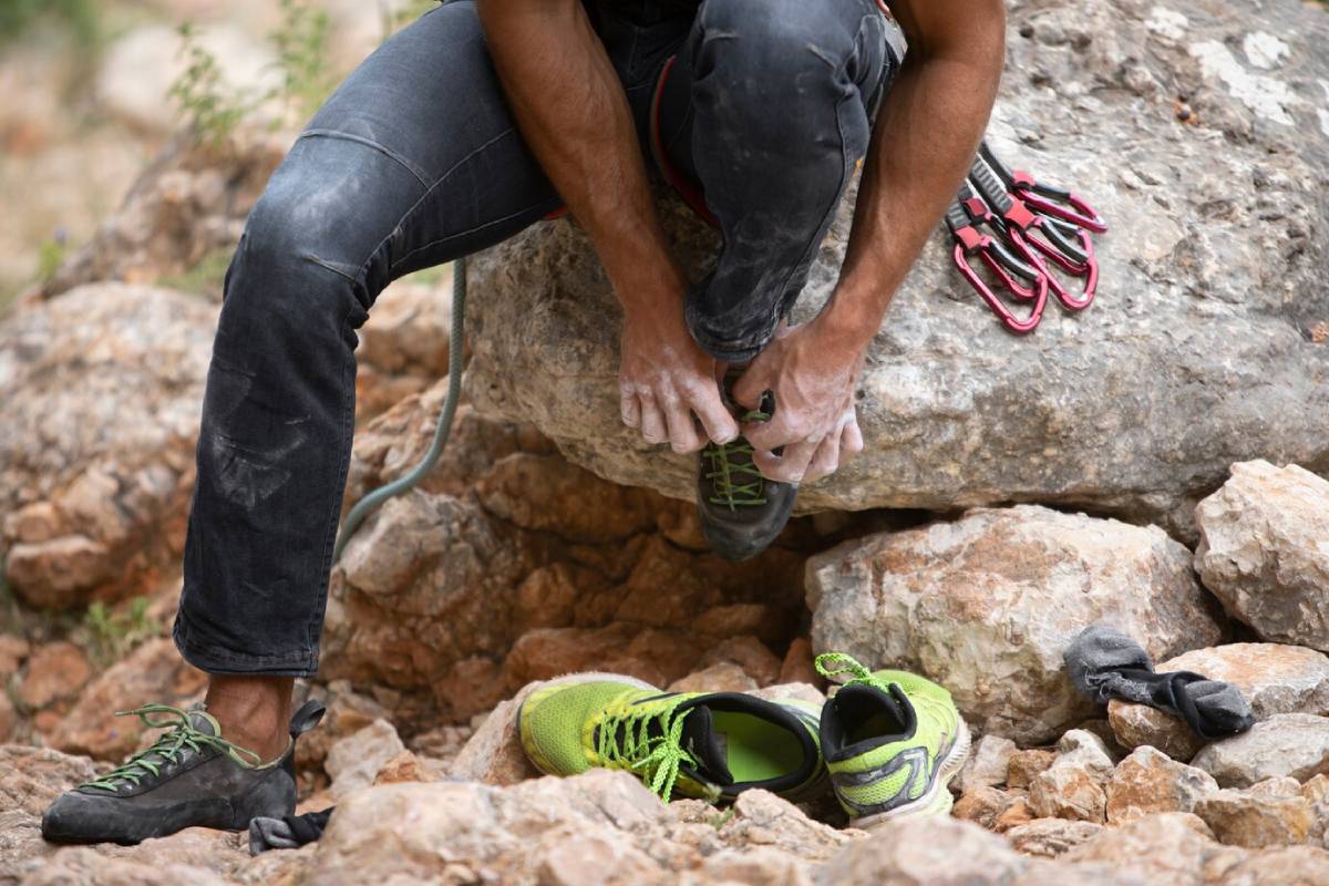  A climber sits on a rock, tying climbing shoes, with a pair of bright green shoes nearby and climbing gear scattered around.