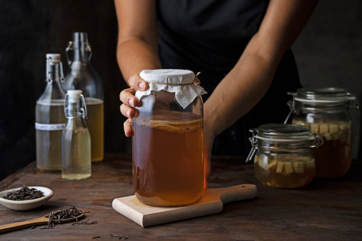 A person holds a large jar of kombucha, surrounded by glass bottles and jars, with loose tea leaves on a wooden board. 