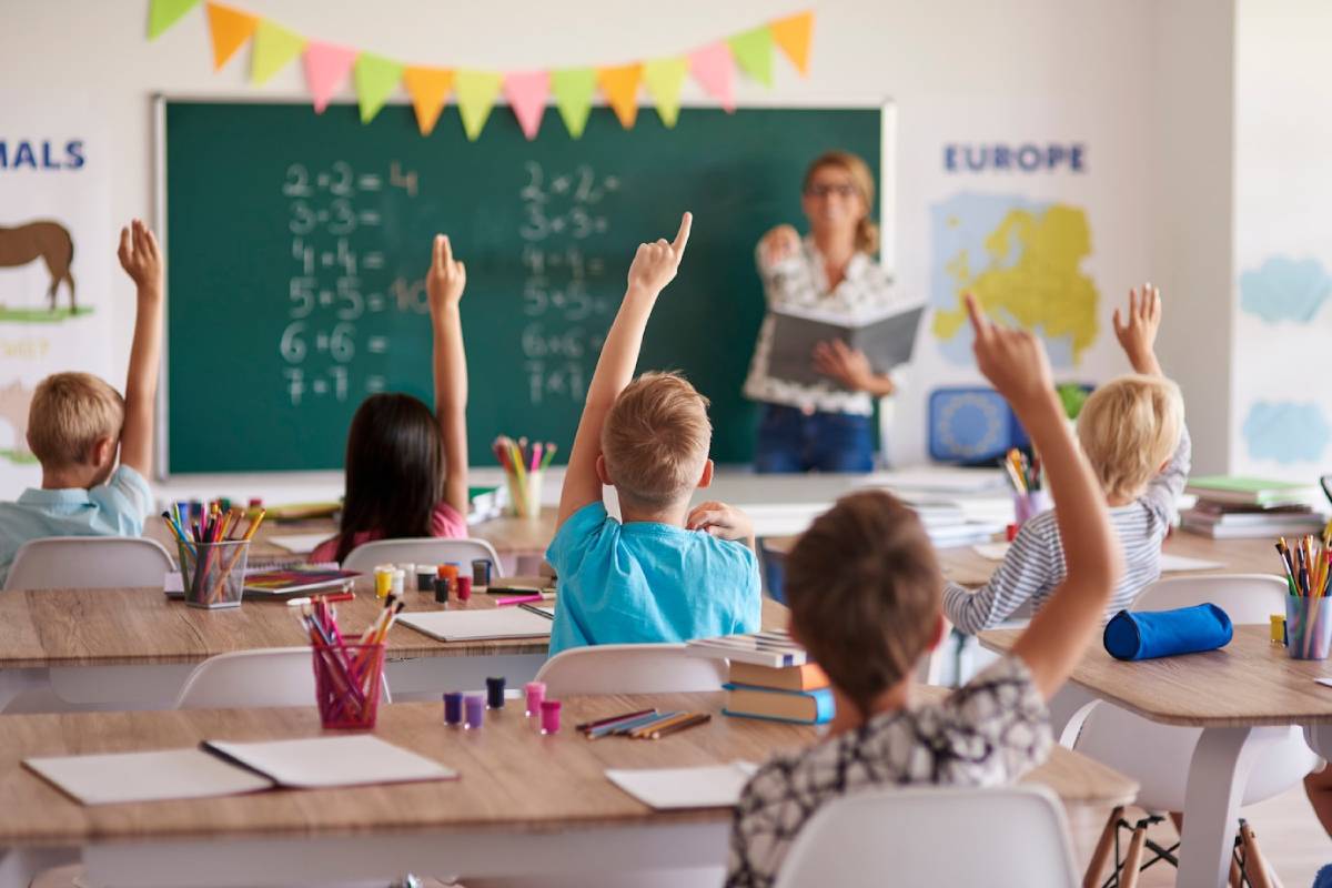  A classroom scene with children raising their hands, seated at tables with colorful supplies, listening attentively to a teacher.