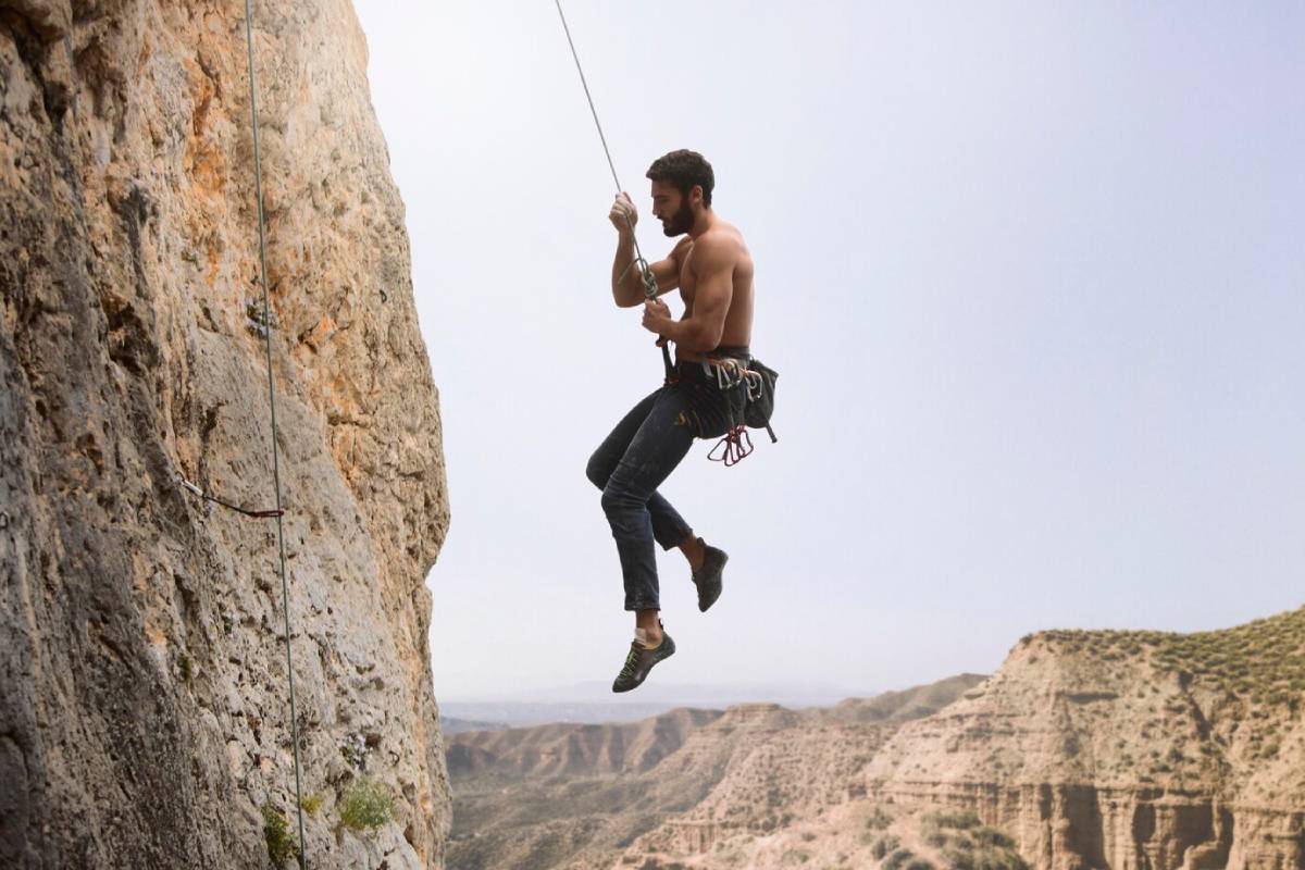  A climber harnessed and suspended from a rock face, surrounded by expansive mountainous terrain and a clear sky.