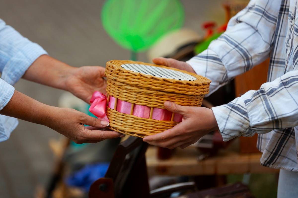  Two hands exchange a woven basket with a pink ribbon, against a backdrop of a vibrant market.