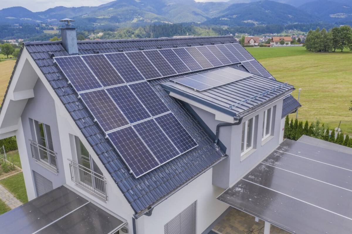 A modern house with solar panels installed on the roof, surrounded by green fields and mountains in the background. 