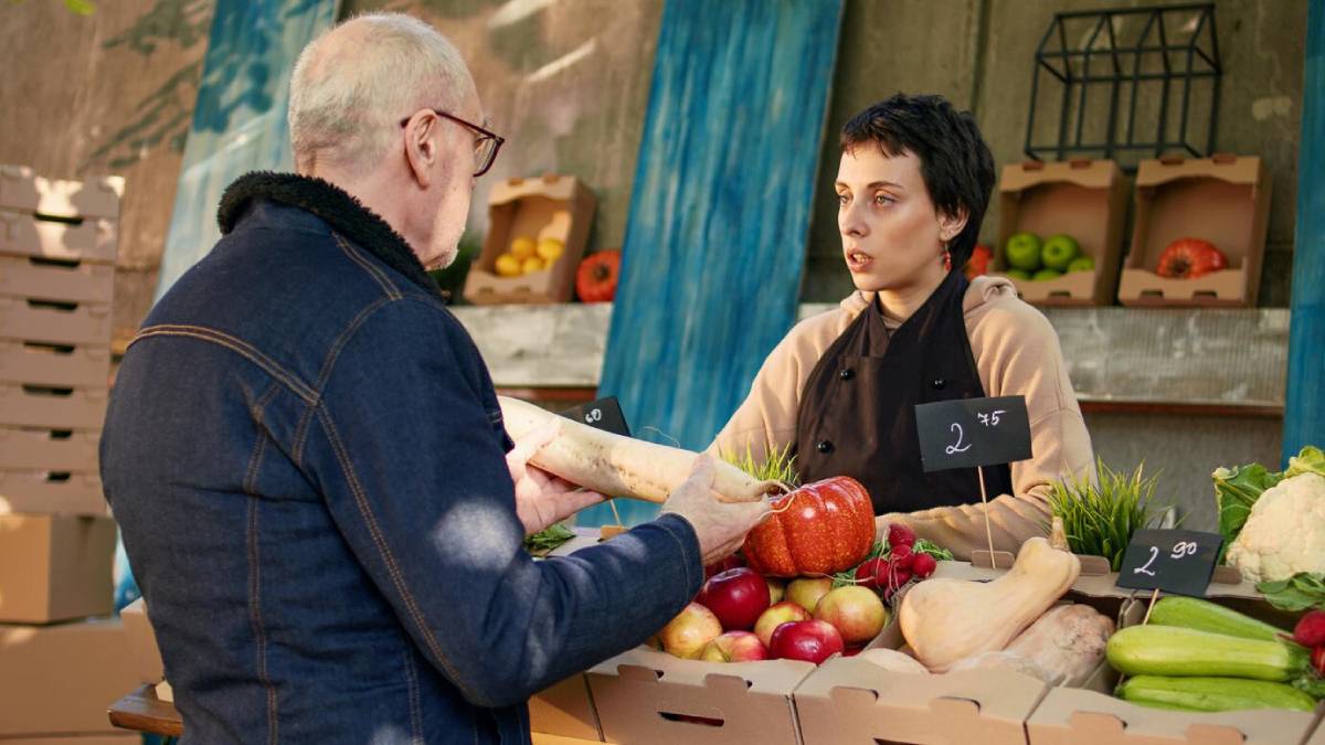 An elderly man talks with a vendor at a market stand full of colorful fruits and vegetables, showcasing fresh produce with price tags. 