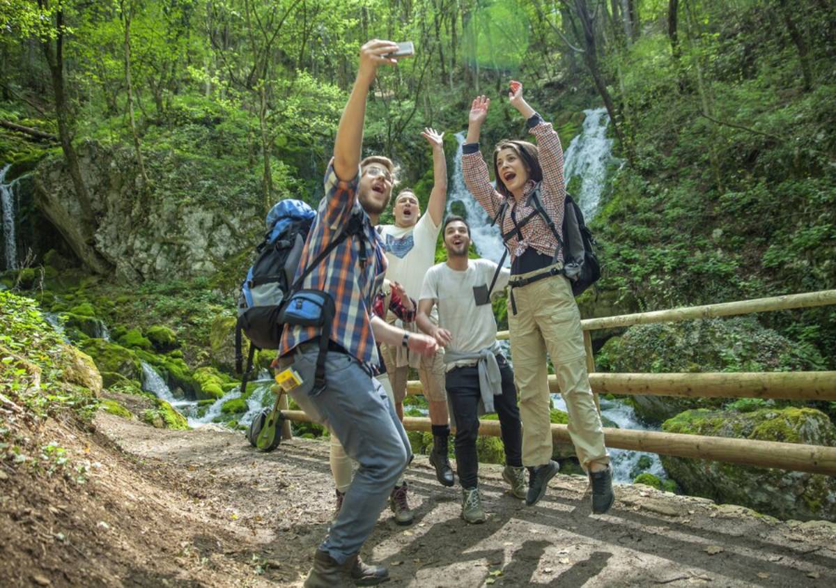 A group of excited hikers jumps and poses for a selfie in a lush forest with a waterfall in the background. 