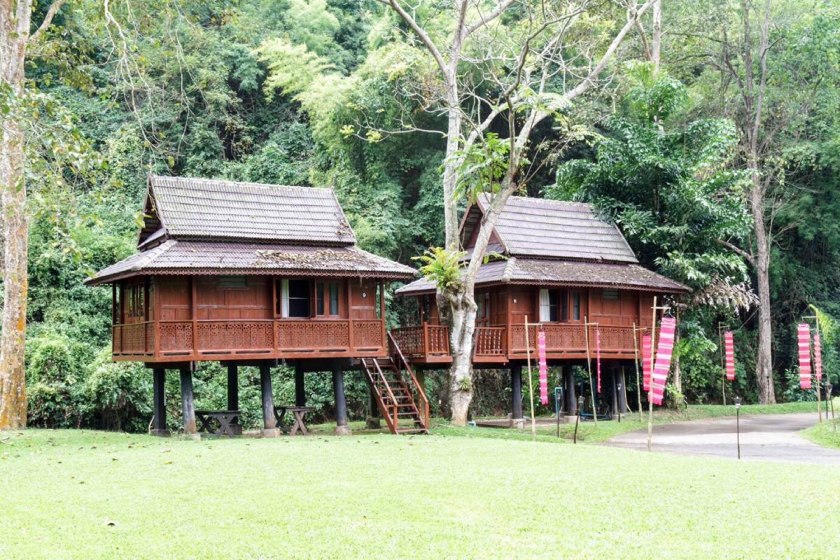 Two traditional wooden houses on stilts surrounded by lush greenery, with decorative banners and a pathway in the background. 