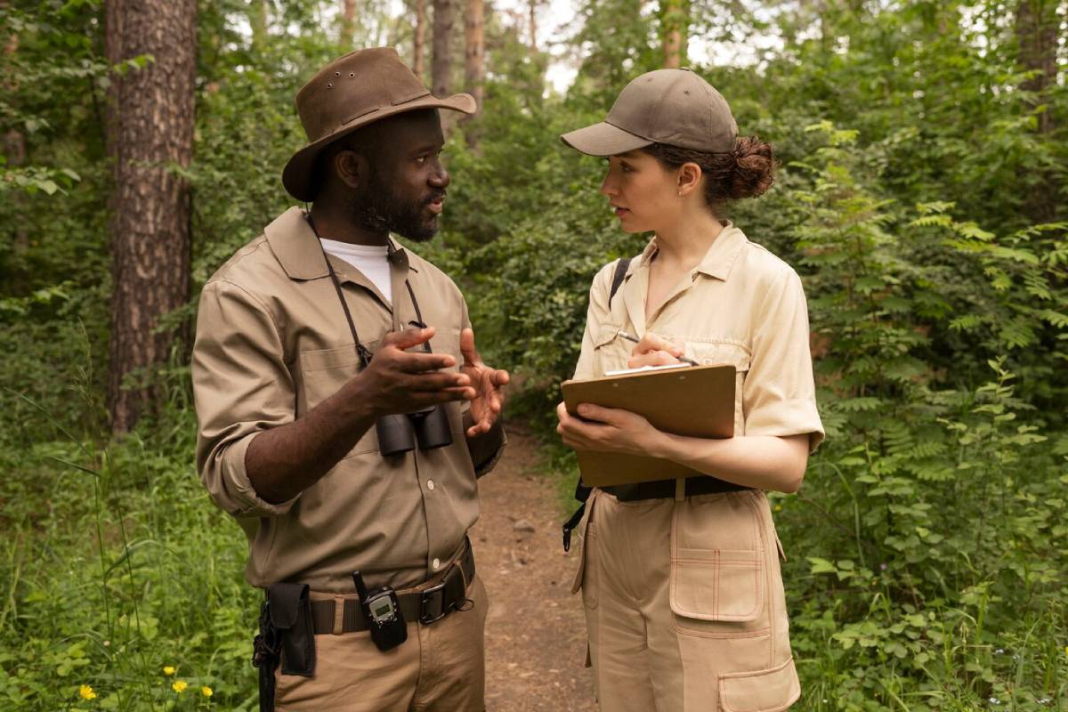 Two wildlife researchers discuss observations in a lush green forest, one holding binoculars and the other taking notes on a clipboard. 