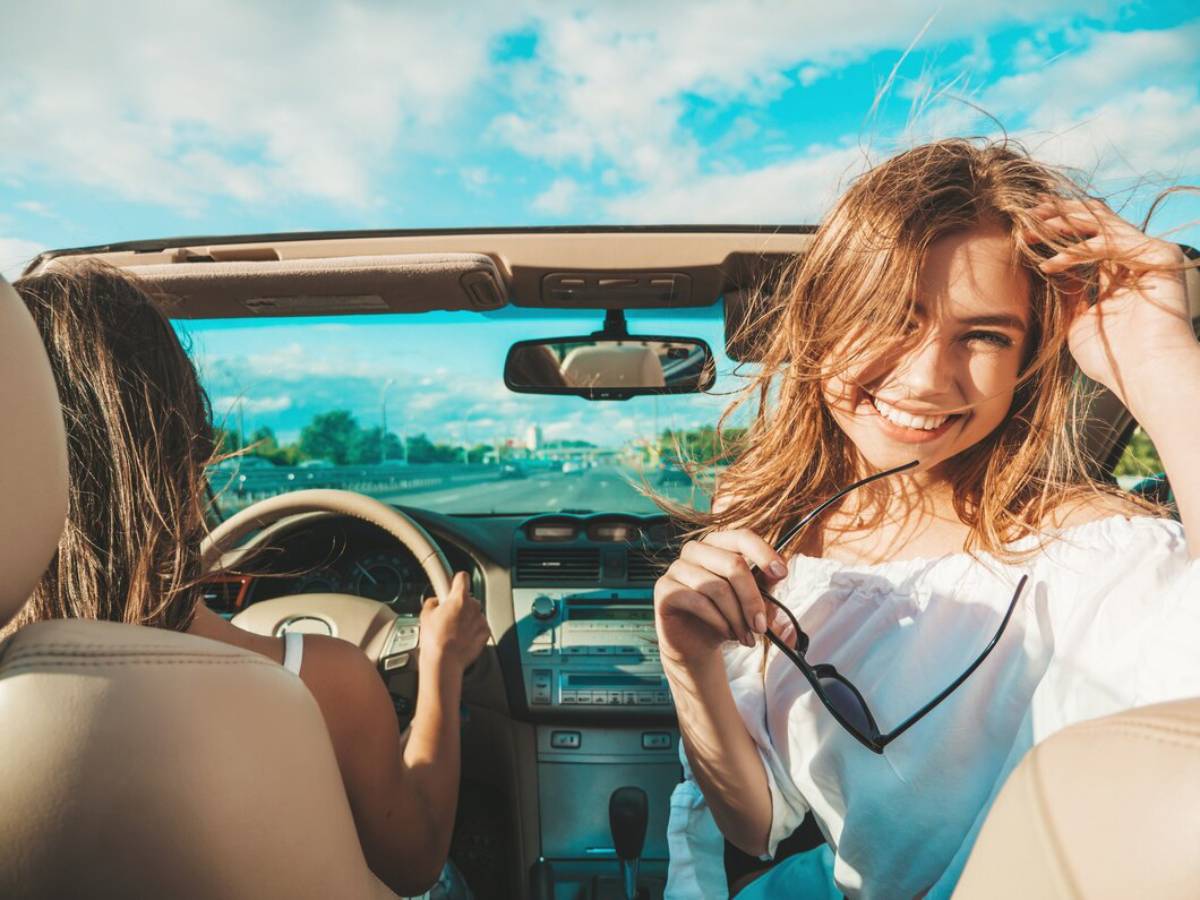  Two women enjoy a sunny car ride with the top down, hair blowing in the wind, against a backdrop of blue skies and open road.