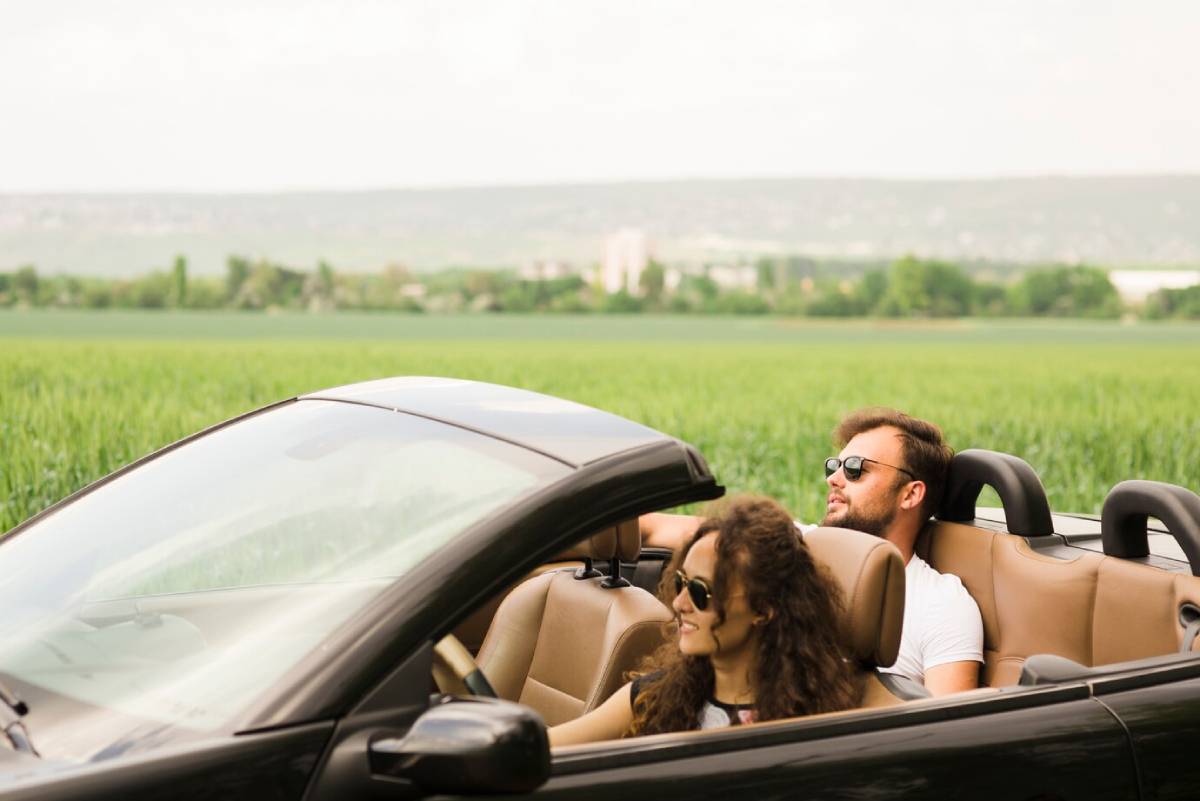 A couple enjoys a ride in a convertible car, with lush green fields stretching out in the background under a cloudy sky. 