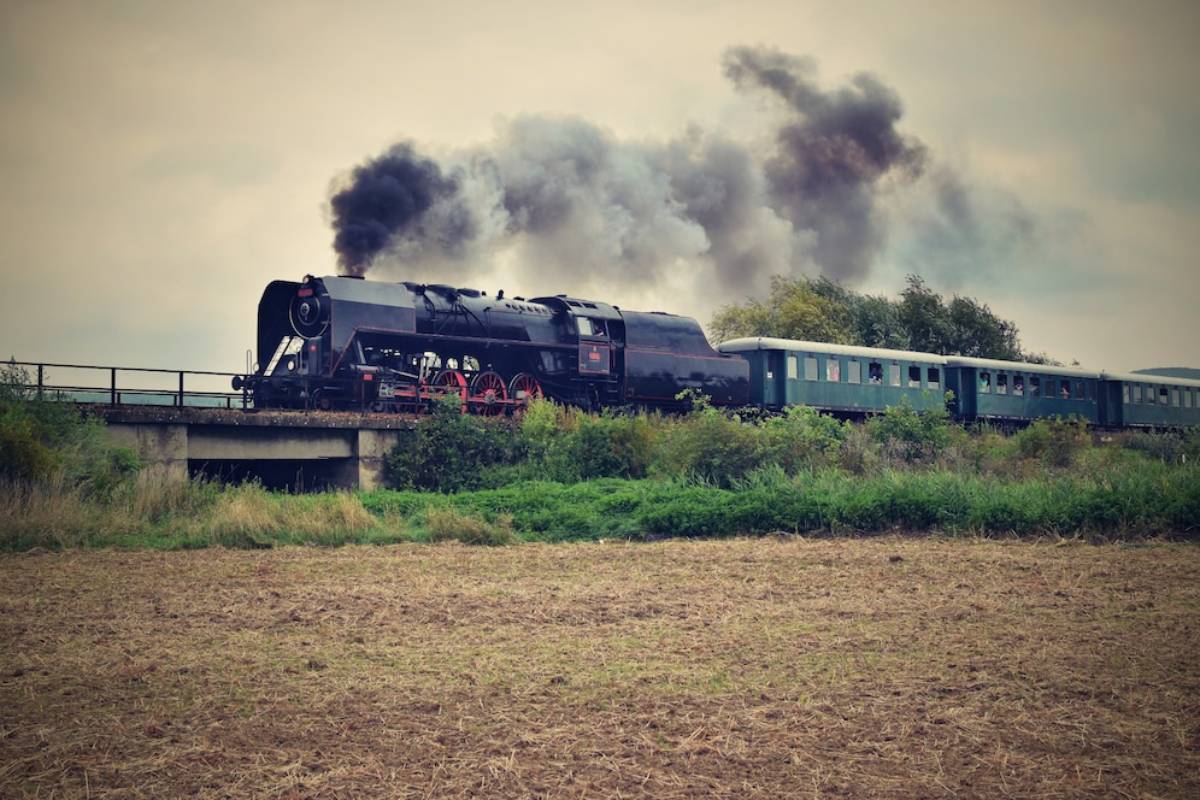 A vintage steam locomotive crosses a bridge, emitting dark smoke, with green passenger cars visible behind it in a rural setting. 