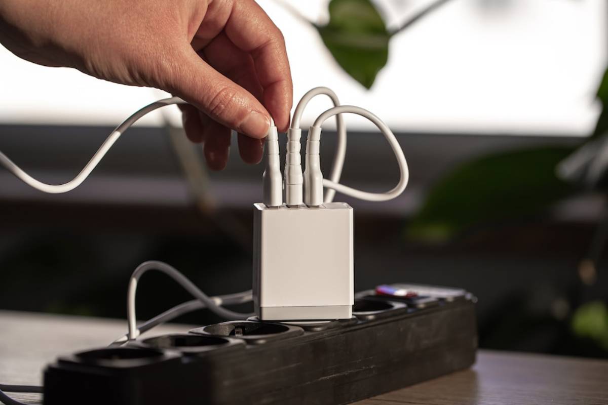 A hand connects multiple charging cables to a white power adapter plugged into a black power strip on a wooden surface. 