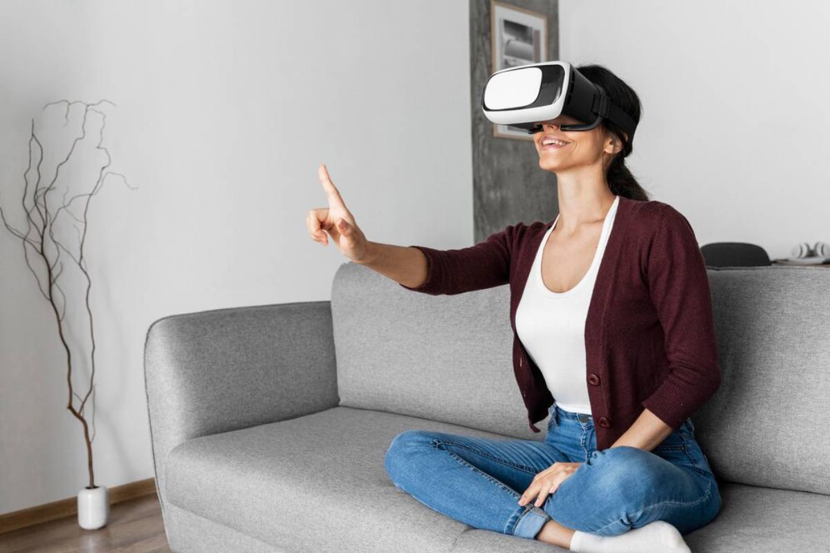 A woman sits on a gray couch, wearing a virtual reality headset, with one hand raised as if interacting with a virtual environment. 