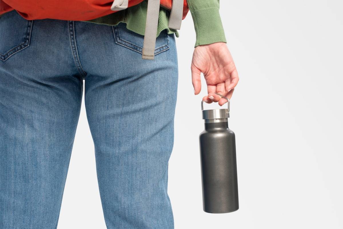  A person in denim jeans holds a dark metal water bottle while wearing an orange backpack and a green sweater.