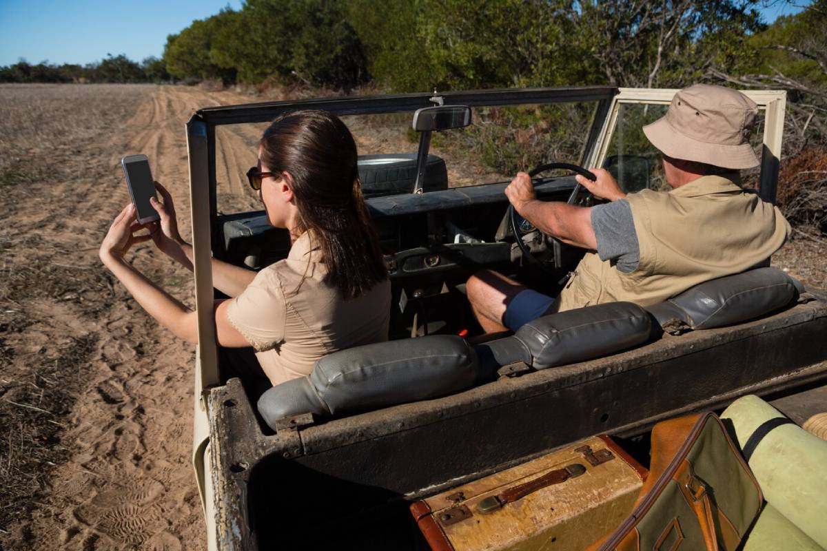  A woman takes a selfie in an open-top vehicle, while a man drives on a dirt path surrounded by greenery.