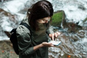 A person wearing a green jacket stands by a flowing stream, focused on using a smartphone. Nature surrounds them with greenery and water.